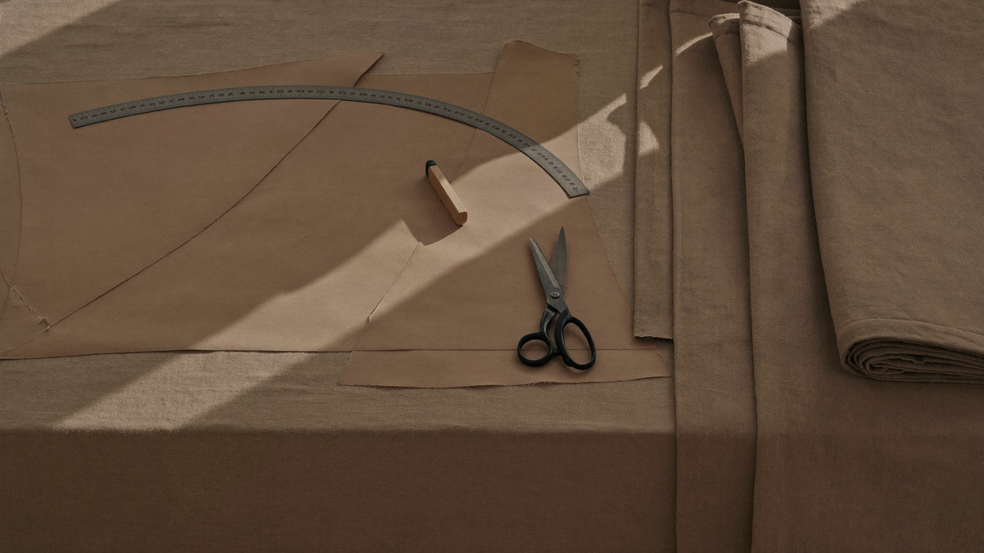 Paper garment pattern pieces, a curved ruler, tailor's chalk and shears on a cutting table with folded linen.