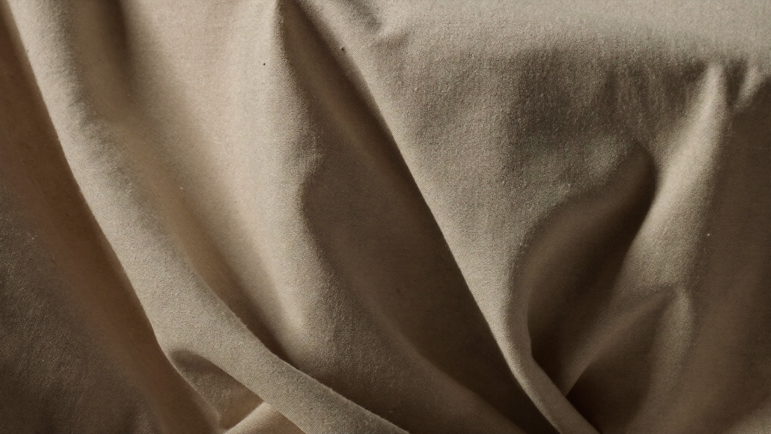 Close up of creased natural linen fabric in warm oatmeal, lit from the side to show the folds.