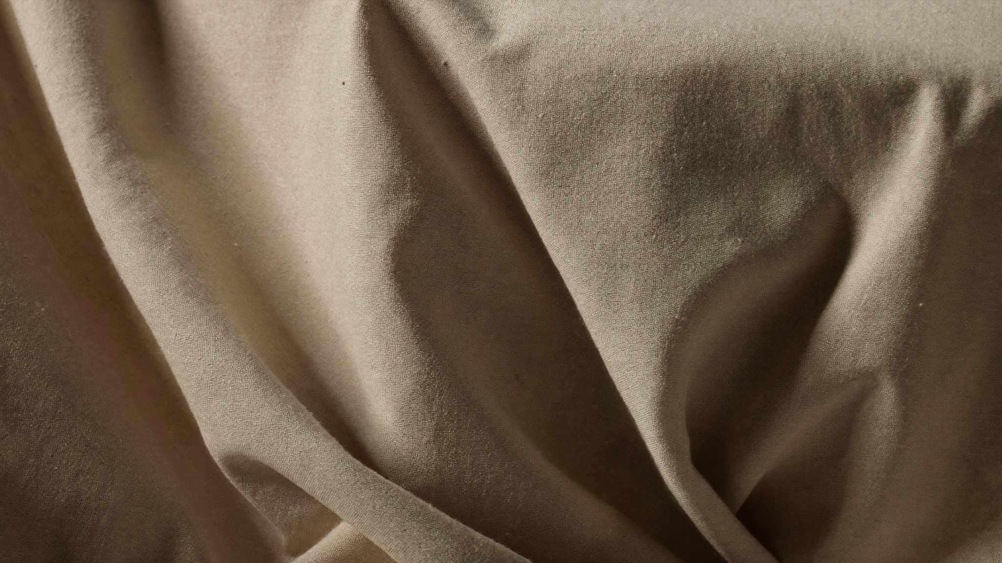 Close up of creased natural linen fabric in warm oatmeal, lit from the side to show the folds.