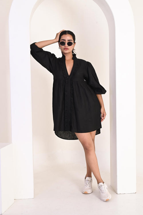May include: A black mini dress with a V-neckline and ruffled trim. The dress has long sleeves and is paired with white sneakers. The model is wearing sunglasses and is leaning against a white archway.