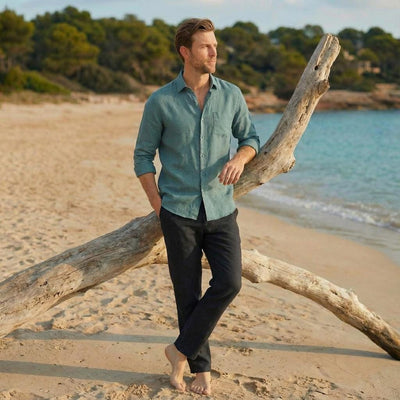 Men's Linen Resort Shirt with Front Pocket: man leaning on driftwood in sage green linen shirt on the beach